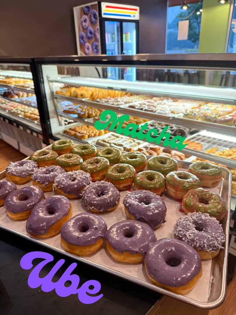 Trays of purple ube donuts topped with shredded coconut next to green matcha donuts with red drizzle, in front of Milkee's filled display cases.