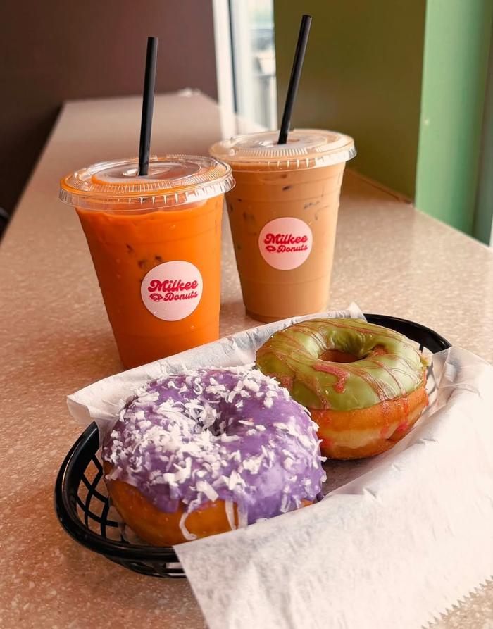 Two Milkee Donuts cups — bright orange Thai tea and an iced coffee — sitting beside a basket holding a purple ube donut under shredded coconut and a matcha donut with red drizzle.